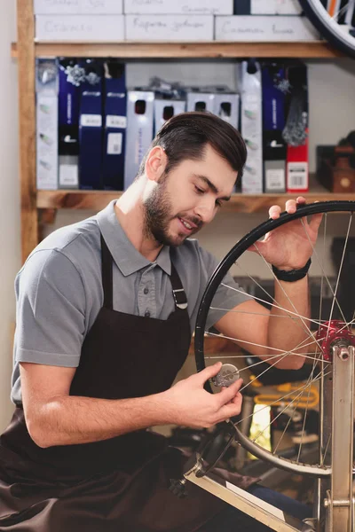 Sorridente giovane in grembiule fissaggio ruota bicicletta in officina — Foto stock