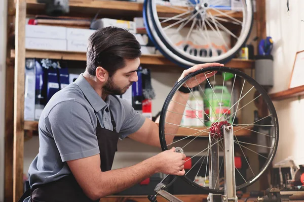 Jeune homme dans tablier réparer roue de vélo dans l'atelier — Photo de stock