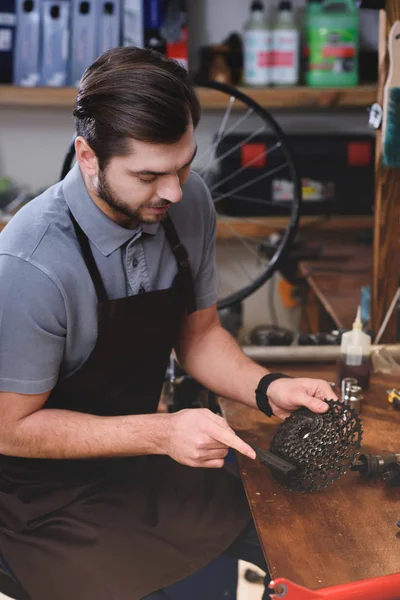 Joven en delantal trabajando con cadena de bicicleta en taller - foto de stock