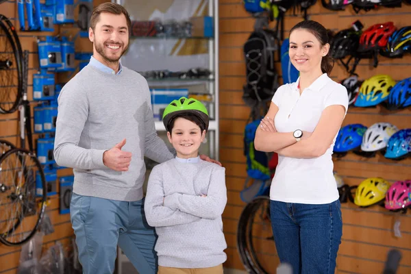 Счастливый отец, милый маленький сын в велосипедном шлеме и продавщица, улыбающаяся на камеру в магазине велосипедов — стоковое фото