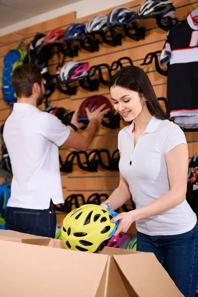 Улыбающиеся молодые рабочие, расставляющие велосипедные шлемы на полках в магазине велосипедов — стоковое фото