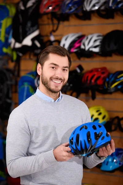 Joven guapo sosteniendo casco de bicicleta y sonriendo a la cámara en el taller — Stock Photo