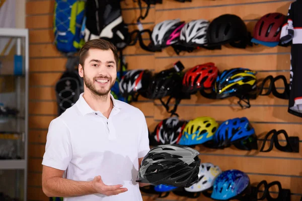 Joven trabajador masculino sosteniendo casco de bicicleta y sonriendo a la cámara en la tienda de bicicletas - foto de stock