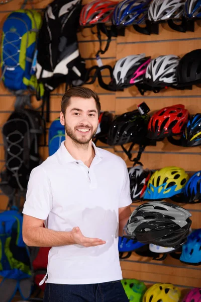 Apuesto joven vendedor sosteniendo casco de bicicleta y sonriendo a la cámara en la tienda de bicicletas - foto de stock