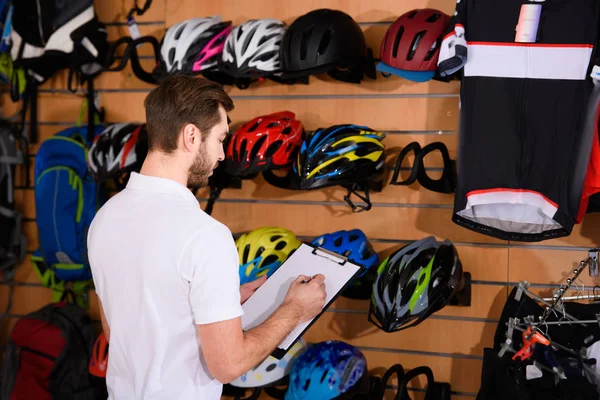 Joven escribiendo en el portapapeles mientras trabajaba en la tienda de bicicletas - foto de stock