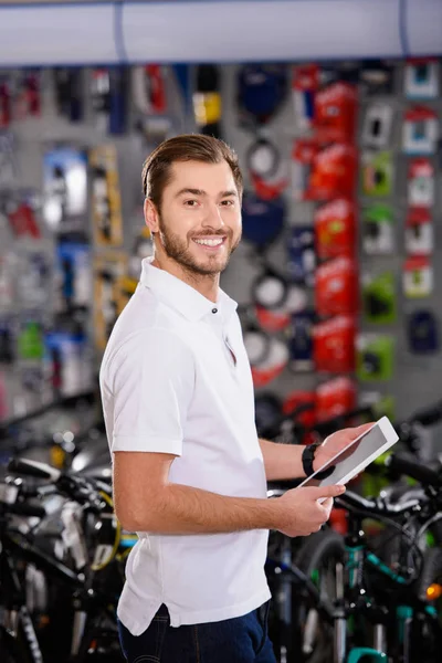 Handsome young man holding digital tablet and smiling at camera in bicycle shop — Stock Photo