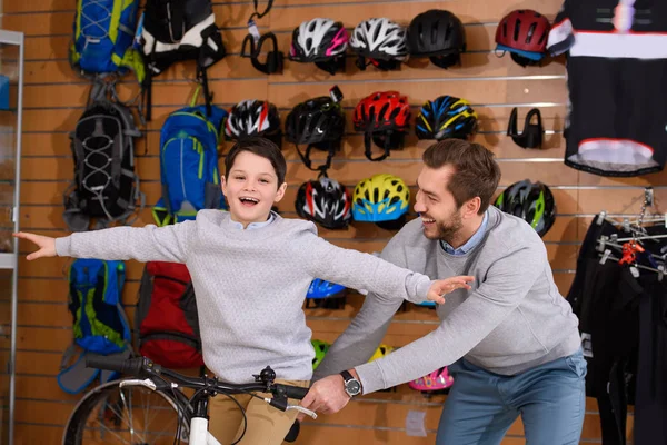 Glücklicher Vater schaut lächelnden kleinen Sohn mit offenen Armen auf Fahrrad im Fahrradladen sitzend an — Stockfoto