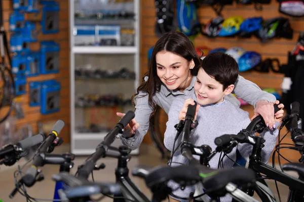 Feliz joven madre con lindo pequeño hijo mirando bicicletas en la tienda de bicicletas - foto de stock