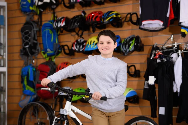 Cute little boy standing with bicycle and smiling at camera in bike shop — Stock Photo