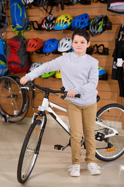 Mignon petit garçon debout avec vélo et regardant la caméra dans le magasin de vélo — Photo de stock