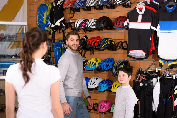 Sonriente padre e hijo eligiendo cascos de bicicleta y mirando a la vendedora en la tienda de bicicletas - foto de stock