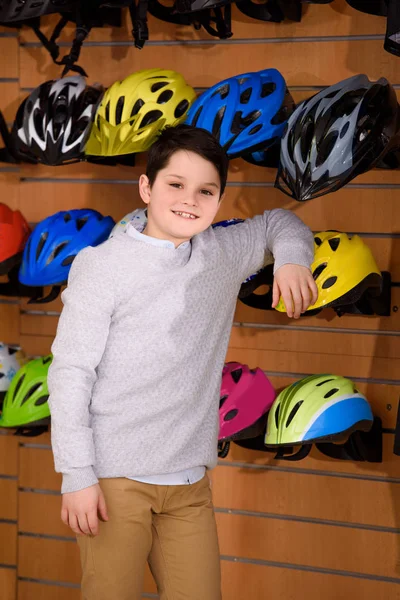 Cute little boy leaning at bicycle helmets and smiling at camera in bike shop — Stock Photo