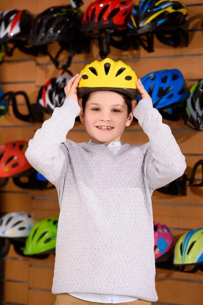 Lindo niño pequeño que se pone el casco de bicicleta y sonriendo a la cámara en la tienda de bicicletas — Stock Photo