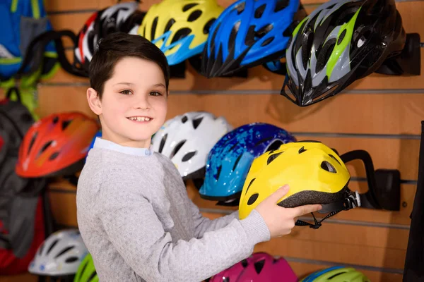 Lindo niño sosteniendo casco de bicicleta y sonriendo a la cámara en la tienda de bicicletas — Stock Photo