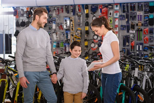 Smiling young woman showing digital tablet to father and son choosing bicycles in bike shop — Stock Photo
