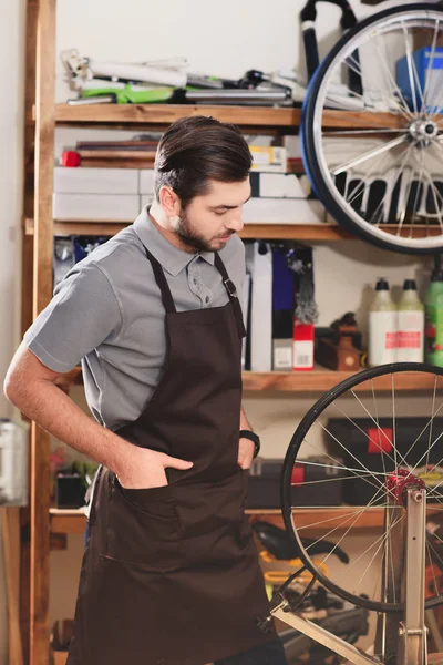 Jovem trabalhador confiante em avental olhando para roda de bicicleta em oficina — Fotografia de Stock