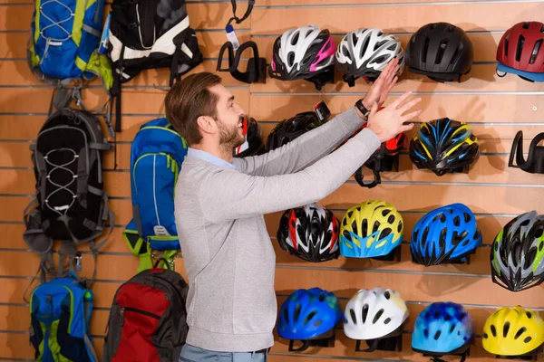 Smiling young man choosing bicycle helmet in bike shop — Stock Photo