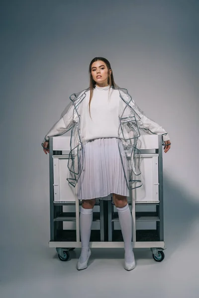 Fashionable woman in white clothing and raincoat posing with collapsible chairs behind on grey background — Stock Photo