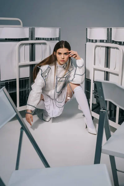 Mulher na moda em roupas brancas posando com cadeiras dobráveis atrás no fundo cinza — Fotografia de Stock