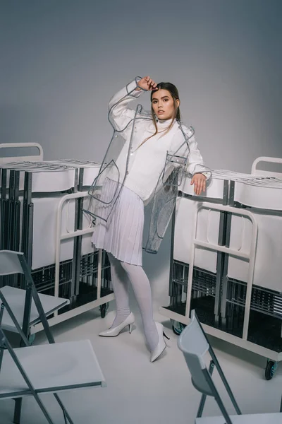 Mulher elegante em roupas brancas e capa de chuva posando com cadeiras dobráveis atrás em fundo cinza — Fotografia de Stock