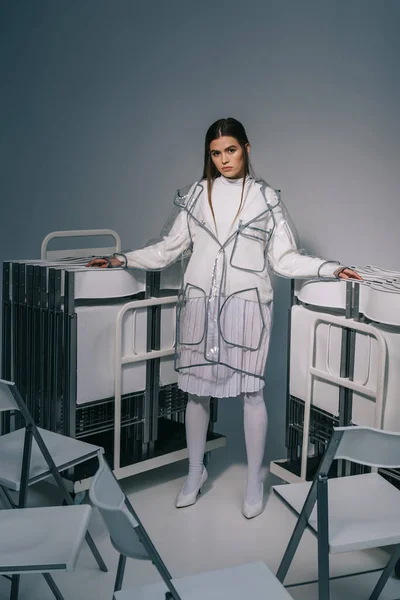 Mulher elegante em roupas brancas e capa de chuva posando com cadeiras dobráveis atrás em fundo cinza — Fotografia de Stock