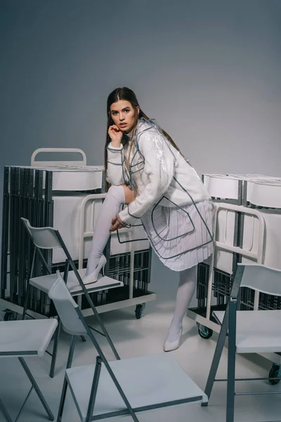 Модная женщина в белой одежде и дождевике позирует со складными стульями сзади на сером фоне — стоковое фото