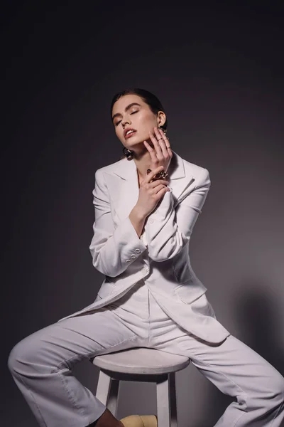 Portrait of fashionable woman in white suit sitting on chair on dark background — Stock Photo