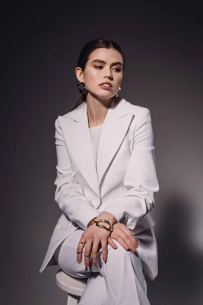 Портрет модной женщины в белом костюме, сидящей на стуле на темном фоне — стоковое фото