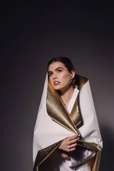 Portrait of fashionable pensive woman in white and golden wrapping paper looking away on dark backdrop — Stock Photo