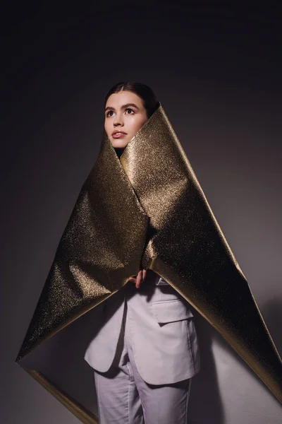 Portrait of fashionable pensive woman in golden wrapping paper looking away on dark backdrop — Stock Photo