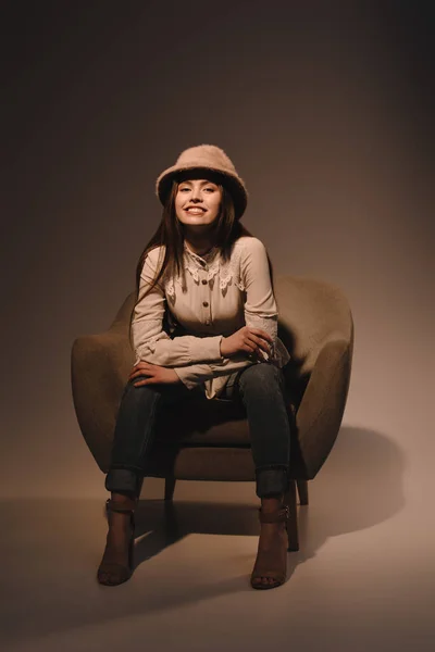 Модная улыбающаяся женщина в шляпе сидит в кресле на темном фоне — стоковое фото