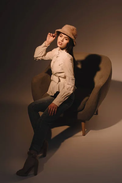 Fashionable pensive woman in hat sitting in armchair on dark backdrop — Stock Photo