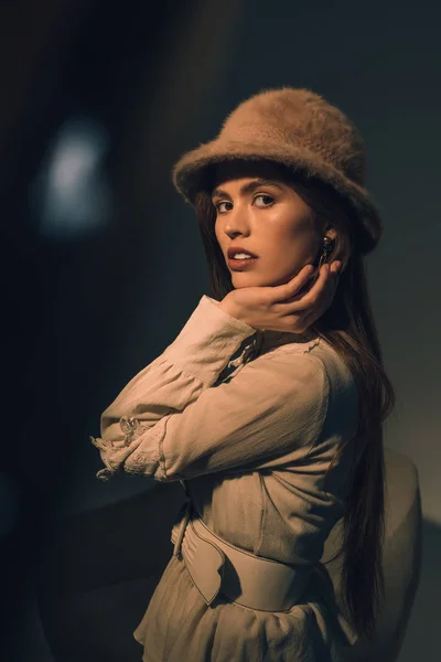 Vista lateral de la hermosa mujer joven en sombrero elegante mirando a la cámara - foto de stock