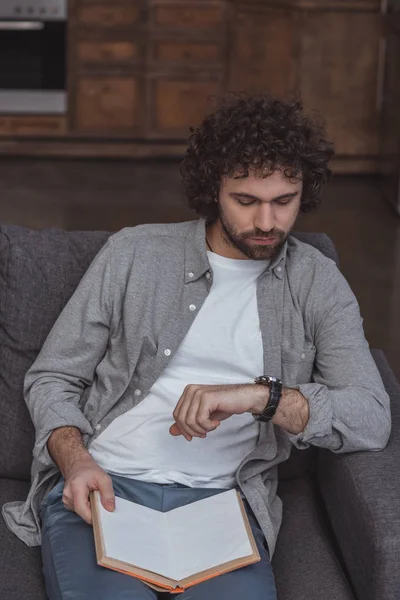 Schöner Mann schaut auf Armbanduhr und hält Buch zu Hause — Stockfoto