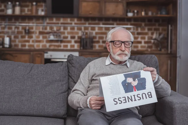 Guapo gris hombre de pelo leyendo periódico de negocios en el sofá en casa - foto de stock