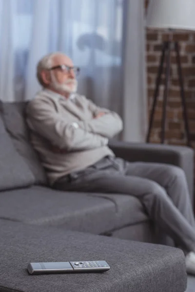 Schöner Mann mit grauen Haaren sitzt auf dem Sofa mit verschränkten Armen und Fernbedienung im Vordergrund — Stockfoto