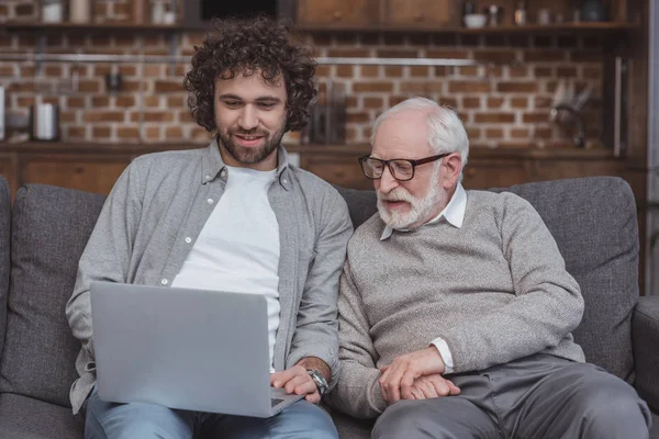 Sorrindo filho adulto e pai sênior usando laptop em casa — Fotografia de Stock