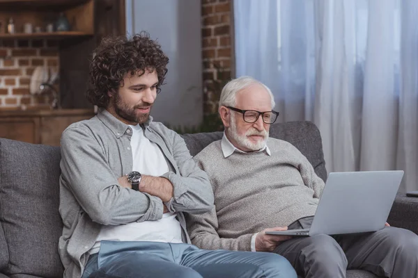 Adult son and senior father using laptop on sofa at home — Stock Photo