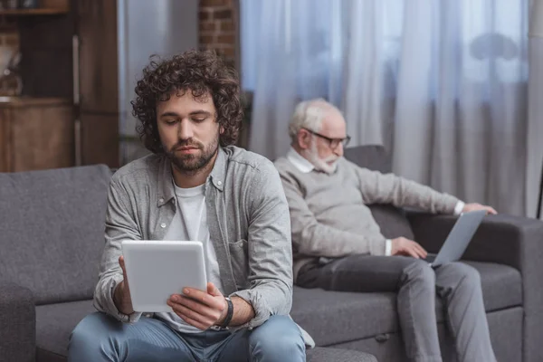 Hijo adulto usando la tableta y padre mayor usando el ordenador portátil en el sofá en casa - foto de stock