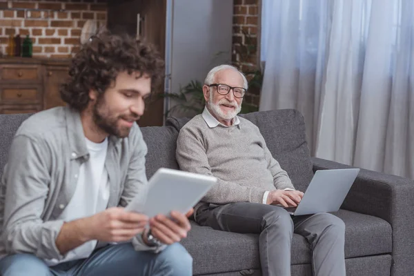 Fils adulte utilisant une tablette et père aîné utilisant un ordinateur portable à la maison — Photo de stock