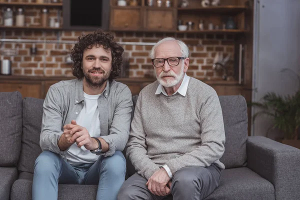 Erwachsener Sohn und älterer Vater sitzen auf Sofa und schauen zu Hause in die Kamera — Stockfoto