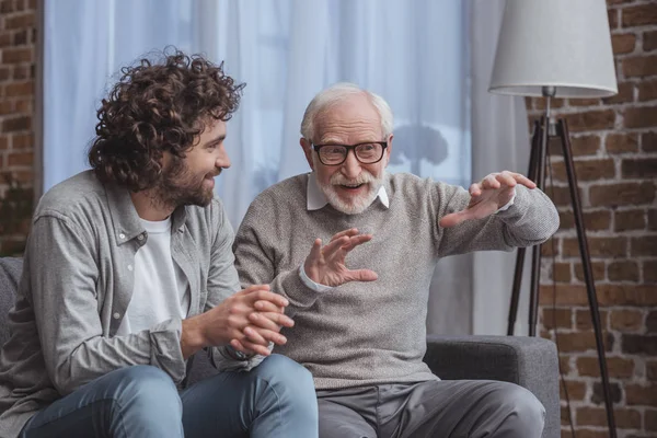 Hijo adulto y padre mayor hablando en el sofá en casa - foto de stock