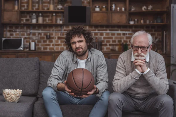 Sad adult son and senior father watching basketball game at home — Stock Photo