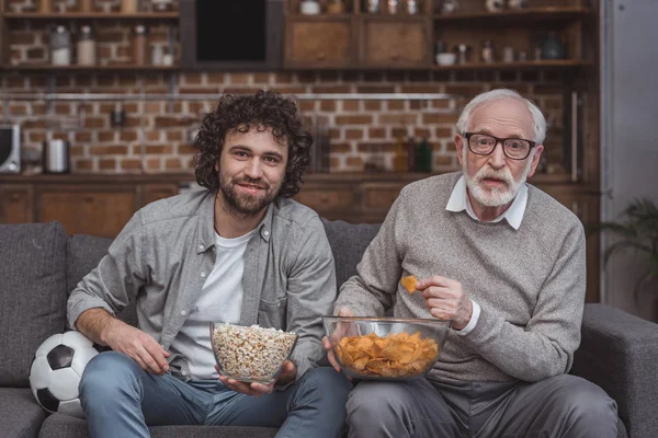 Adult son and senior father watching football game with popcorn and chips at home — Stock Photo