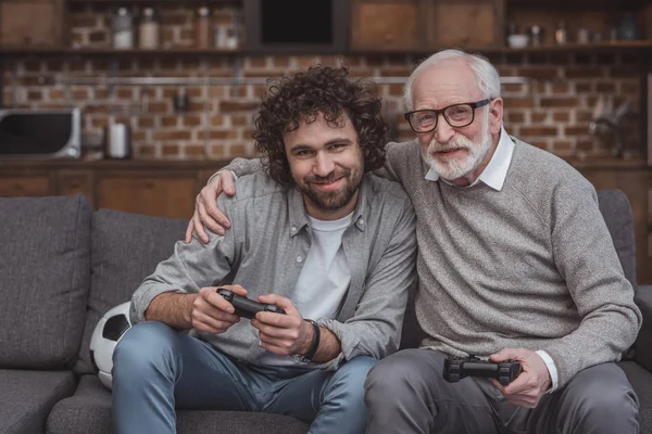 Père aîné câlin fils adulte tout en jouant à un jeu vidéo à la maison — Photo de stock