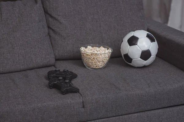 Tazón con palomitas de maíz, pelota de fútbol y gamepads en el sofá - foto de stock