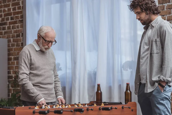 Hijo adulto y padre mayor jugando futbolín con cerveza en casa - foto de stock