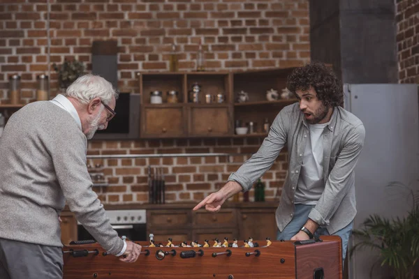 Überraschter erwachsener Sohn zeigt auf Tischkicker, während er mit Senior-Vater zu Hause spielt — Stockfoto