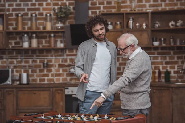 Filho adulto e pai sênior apontando para o futebol de mesa em casa — Fotografia de Stock