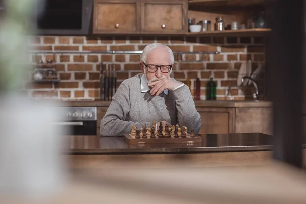 Вдумчивый красивый седой мужчина думает и смотрит на шахматную доску дома — стоковое фото